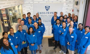 A large group of scientists and visitors wearing blue lab coats gathers in front of the Artificial Intelligence for Materials Design Laboratory.