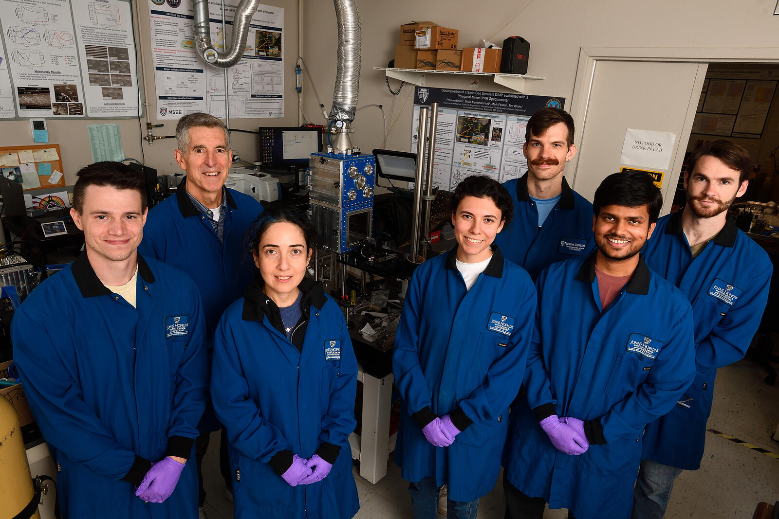Tim Weihs and his students and postdocs stand in the lab, all wearing blue lab coats and purple gloves.